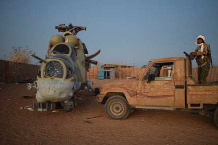 An FLA fighter stands on the flatbed of a pick-up truck next to a damaged helicopter in a dusty desert compound with a low fence. Both the truck and the helicopter look old and hard-used. The fighter wears camouflage and has a white scarf wrapped around his head and a gun over his shoulder.
