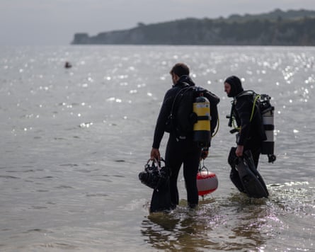 Two men in diving gear and wetsuits walk into the water.