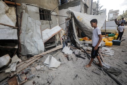A boy stands staring in dust and rubble beside the collapsed wall of a building