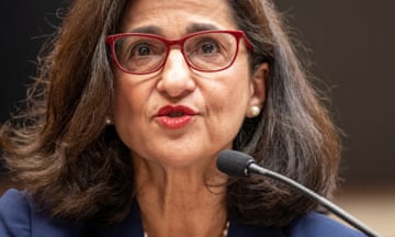 Middle-aged white woman with thick dark hair and red-framed glasses, wearing red lipstick, speaks into a microphone.