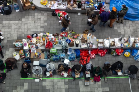 people sit in a line eating meals, next to a long table full of various food and supplies