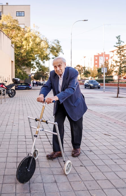 Older man walks along a pavement using a three-wheeled walker.