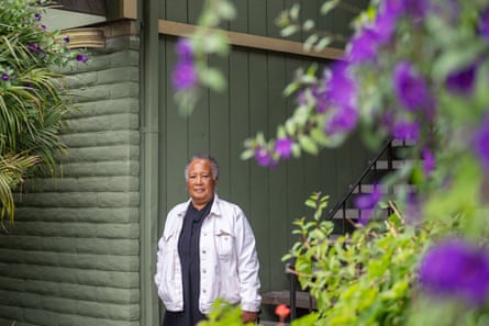 A woman in a white long-sleeved denim jacket, leaning against an olive-painted wall, with purple flowers blurry in the foreground.