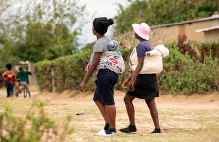 Two young women stroll along a residential dirt street with babies strapped to their backs.