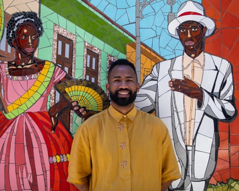 A man in a yellow shirt stands against a mosaic depicting a of a man in a white suit and a lady is a pink dress
