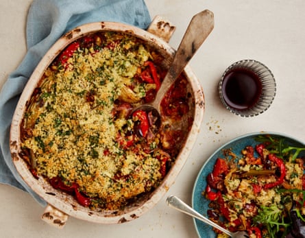A casserole dish of fennel and red pepper bake with a wooden spoon in it, supported on a cloth napkin, next to a glass of red wine and a plateful of the bake with a fork