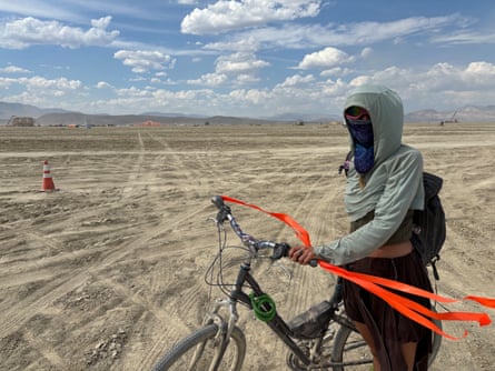 A woman wearing a bandana around her face holds a bike in the desert