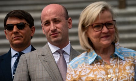 A line of three people, with two middle-aged white men in suits on the left and one middle-aged white woman with blond hair, glasses and a blue and yellow blouse on the left.