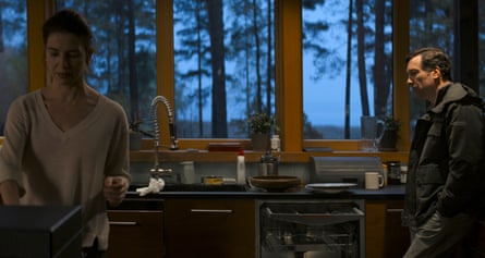 A couple in a kitchen with the woman with her back to the man, who is standing in his jacket