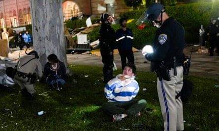 At night, a young man in a bright white and blue striped shirt, his hands tied behind his back, sits crosslegged on a green lawn as a police officer writes something with a flashlight, and beyond him, another police officer does the same with another student.