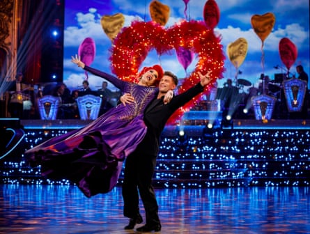 A drag queen in a red wig and purple gown is swept off her feet while dancing with a man dressed in all black.