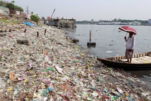 A man holds a red parasol as he inspects a huge pile of waste including plastic covering the banks of the River Buriganga in Dhaka, Bangladesh