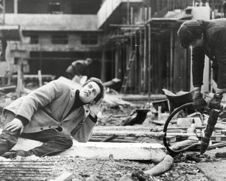 A man wearing a jacket crouches with ear to the ground amid roadworks, as a man operates a jackhammer nearby