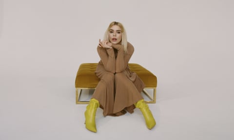 Billie Piper sitting on a rich velvet stool wearing a brown dress and bright yellow leather boots