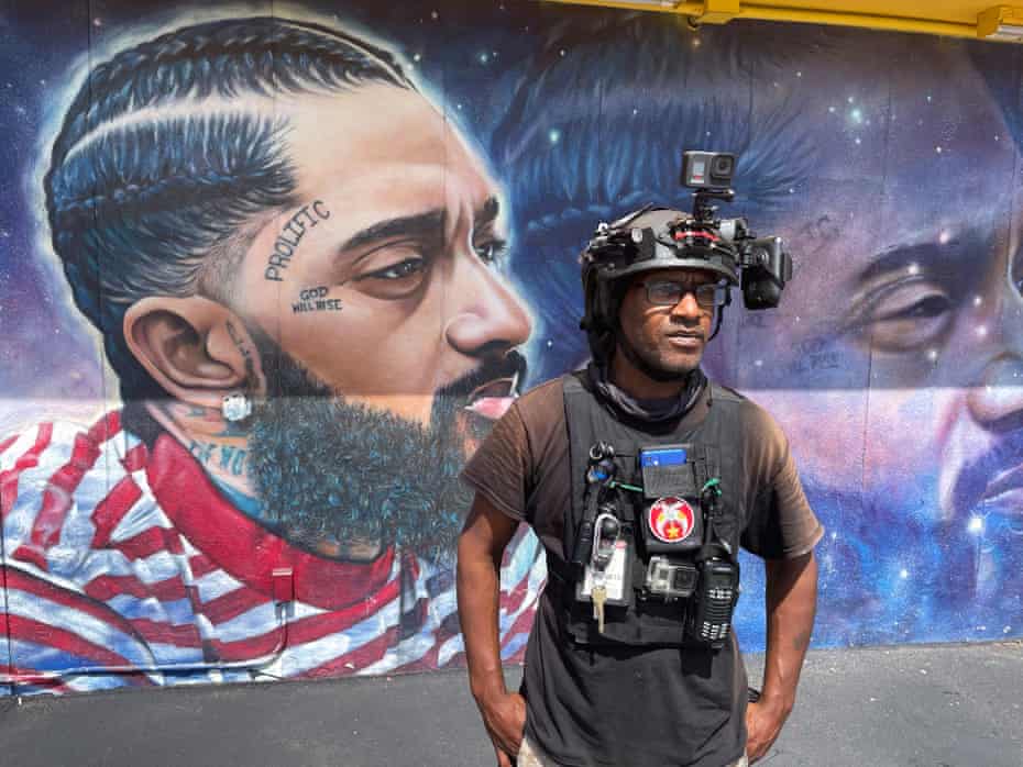 A man wearing video gear stands in front of a mural of a Black man
