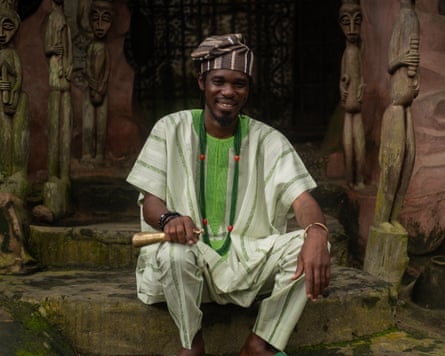 A young African man in traditional dress sitting in a shrine surrounded by wooden fetishes