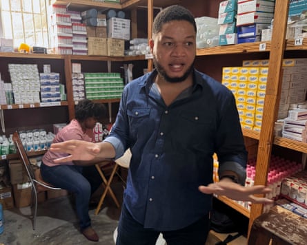 A man, mid-conversation, stands in a storeroom containing shelves stacking with boxes of medical products