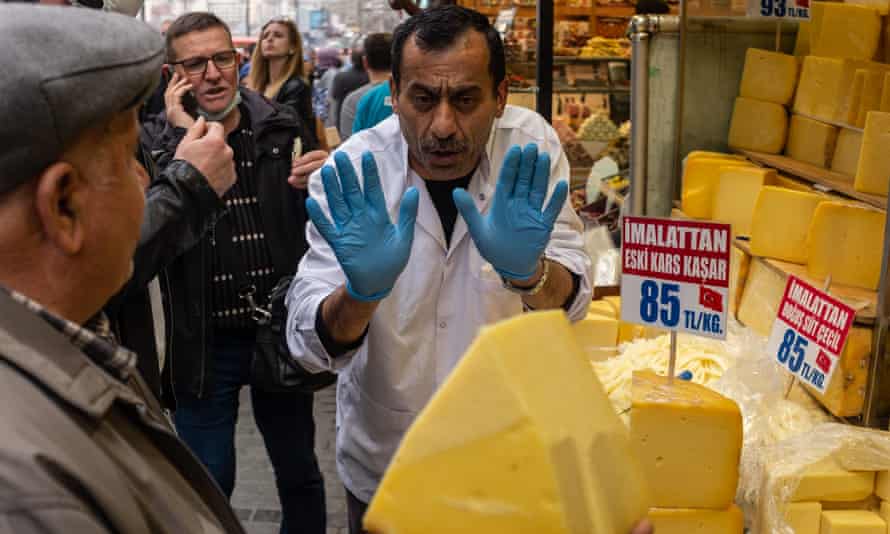 A vendor wearing hygienic rubber gloves gestures with both hands to a customer holding up a large lump of cheese at a market stall