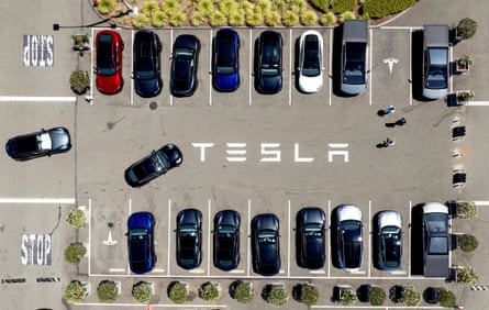 Overhead view of Tesla vehicles parked in two lines and with two manoeuvring. The brand-name Tesla is stencilled in white in the centre of the parking area and logos are seen in the empty spaces.