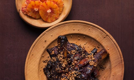 Dark brown tamarind pork ribs with a covering of seeds on a plate and a dish of orange slices next to it.