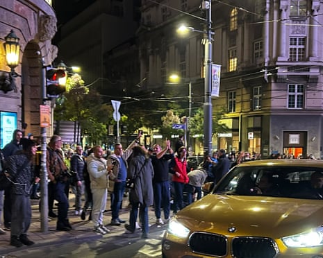 A crowd celebrates on a Belgrade street corner at night as a car passes by