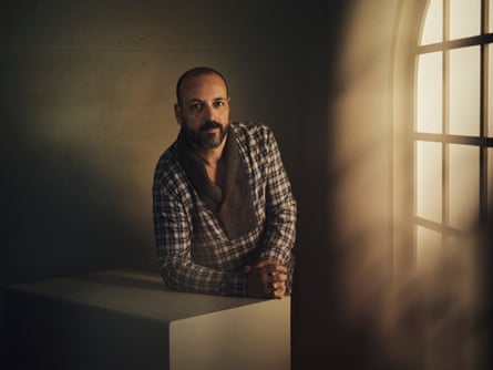 A man in shown in a dimly lit room next to an arched window where light is coming in from.