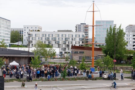 Wide-shot from across road of mast and crowd with buildings behind