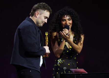 Sam Fender holds a trophy as Olivia Dean speaks into a microphone on stage