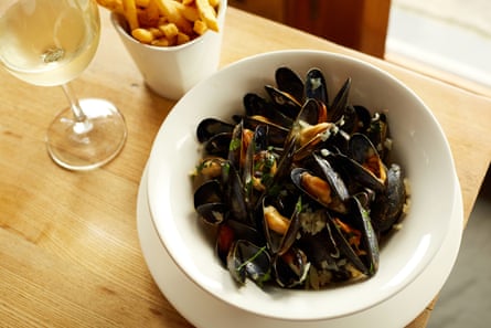 A bowl of mussels in broth with a glass of white wine and a container of chips next to it.
