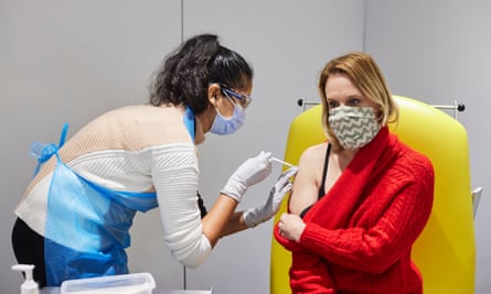 Latina woman wearing facemask leans over white woman holding red sweater down to inject upper arm.
