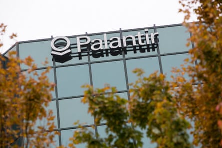A glass-fronted office building occupied by Palantir in Virginia, US, with the company’s logo across the top floor’s windows. There are trees with orange-brown leaves in the foreground.