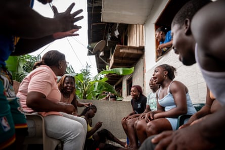 A group of people sit together on chairs and chat, one woman leans out a window