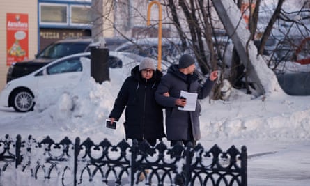 Lyudmila Navalnaya, the mother of Alexei Navalny, and his lawyer, Vasily Dubkov: they are both dressed in thick coats and woollen hats, and are walking behind iron railings; thick snow is seen in the background