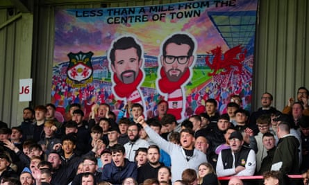 Wrexham supporters cheer in the stand in front of a large banner that depicts the club’s stadium and its owners in cartoon form wearing Wrexham scarves