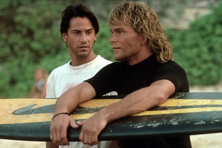 Keanu Reeves next to Patrick Swayze who holds a surf board.
