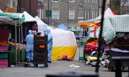 Officers in blue forensic suits speak to a man amid market stalls