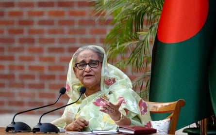 Sheikh Hasina sitting at a table with microphones on it and a Bangladesh flag behind