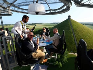 A waiter in a safety harness serves wine to diners with a view of the countryside below them