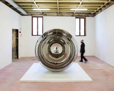 A large spherical metallic sculpture with concentric circular rings sits on a white plinth in a gallery space.