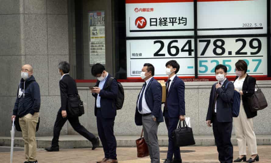People wearing protective masks stand in front of an electronic stock board showing Japan's Nikkei 225 index 