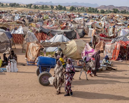 Two African women and a donkey cart pass a large number of makeshift tents on a sandy plain