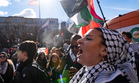 A crowd of people carrying signs and Palestinian flags march, with a woman in the foreground wearing a keffiyeh on her head appearing to chant or yell.