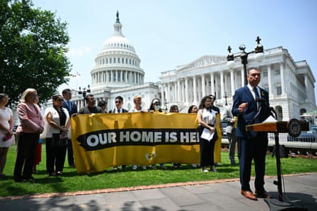 a man speaks into a microphone as a group of people behind him hold a banner that reads ‘our home is here’