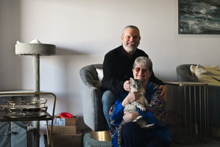 Older white couple sit together in an armchair in a tidy, spare room, smiling, she holding a cat.