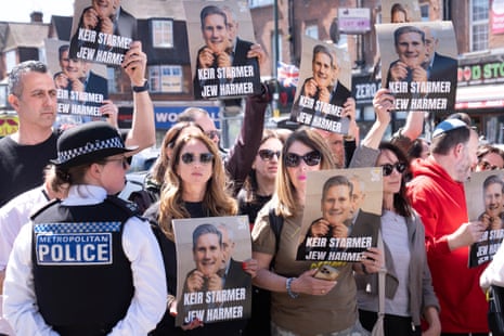 A police officer stands in front of a crowd of protesters holding placards that read 'Keir Starmer, Jew harmer'.