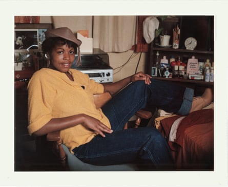 A woman sitting sideways on a plastic chair in her cell, wearing a straw hat. The cell has a record player, knick-knacks and toiletries in it.