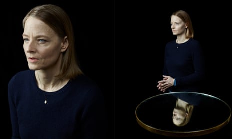 Jodie Foster in double, with the one behind standing next to a glass reflecting her image. The background is black