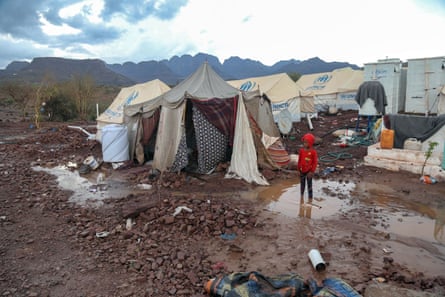 A child stands in the mud wearing a red hoody outside makeshift tents in a desolate scene