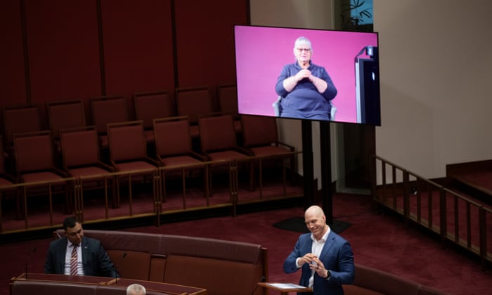 David Pocock stands and speaks in the Senate with an Auslan interpreter being broadcast on a screen behind him. The interpreter on the screen is sitting in a chair in front of a pink background