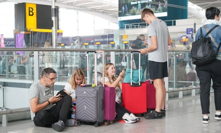 passengers wait at Heathrow airport: two young women and a young man sit on the floor by their wheelie cases in front of a glass barrier, looking at their phones. A fourth young man is standing with them. The four are all causally dressed in white or grey T-shirts and black shorts or jeans with trainers; their cases are grey, dark pink and red, and one of the women has a mint-green handbag on top of her red case. Another person in a denim jacket and black jeans with a black rucksack is seen from behind walking past.